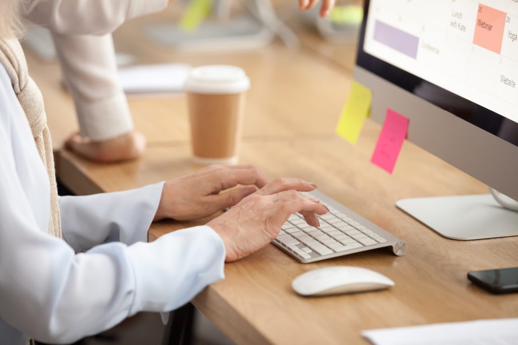 older-female-worker-typing-on-keyword-next-to-colleague-and-takeaway-coffee-cup